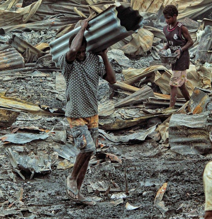 Two people carrying metal sheets through rubble, showing hard work and effort amidst debris.