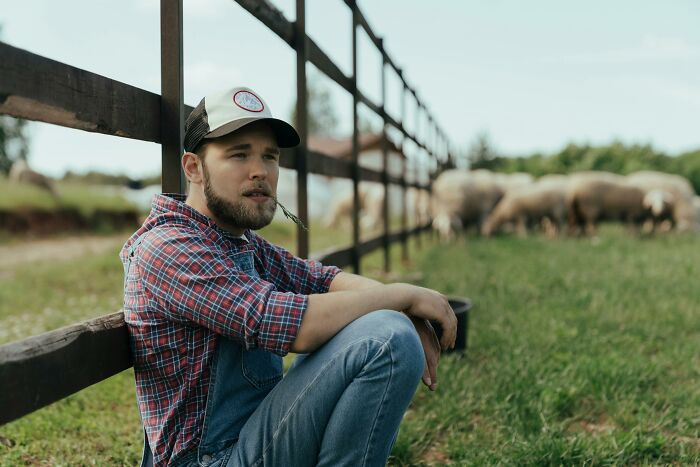 Man in plaid shirt and cap sitting by a fence, thoughtful expression, showcasing signs of subtle intelligence.
