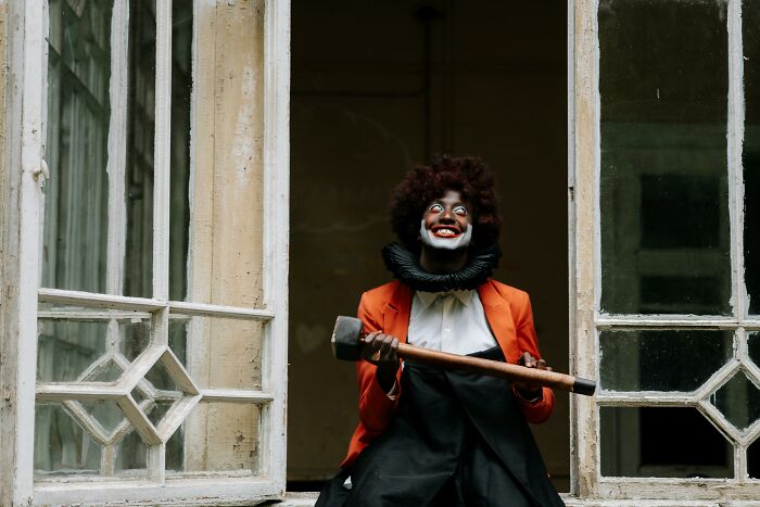 A person in a clown costume holding an axe, sitting in a window frame, evokes eerie wild theories.