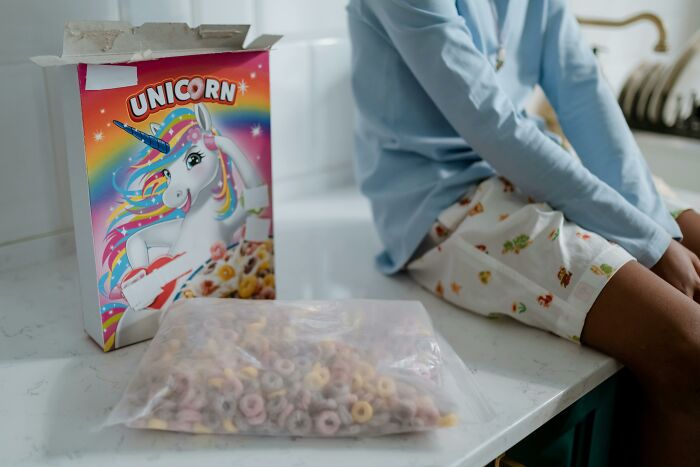 A box of nostalgic discontinued unicorn cereal next to a seated child in a kitchen.