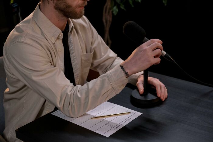Person with a microphone, wearing a beige shirt, sitting at a table with a compliment questionnaire and a pen.