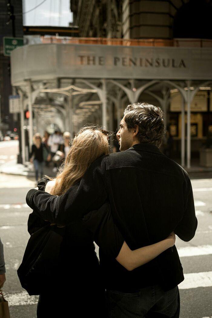 Couple standing together on a city street, sharing a moment near a building with "The Peninsula" sign in the background.