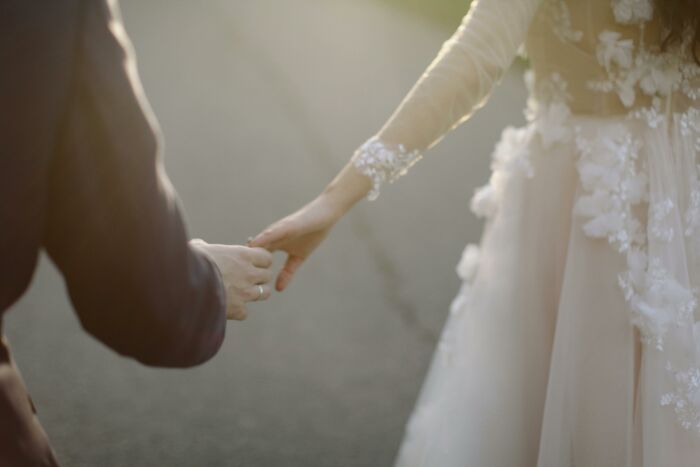 Close-up of a couple holding hands outdoors, with focus on intricate lace details of the woman's dress.