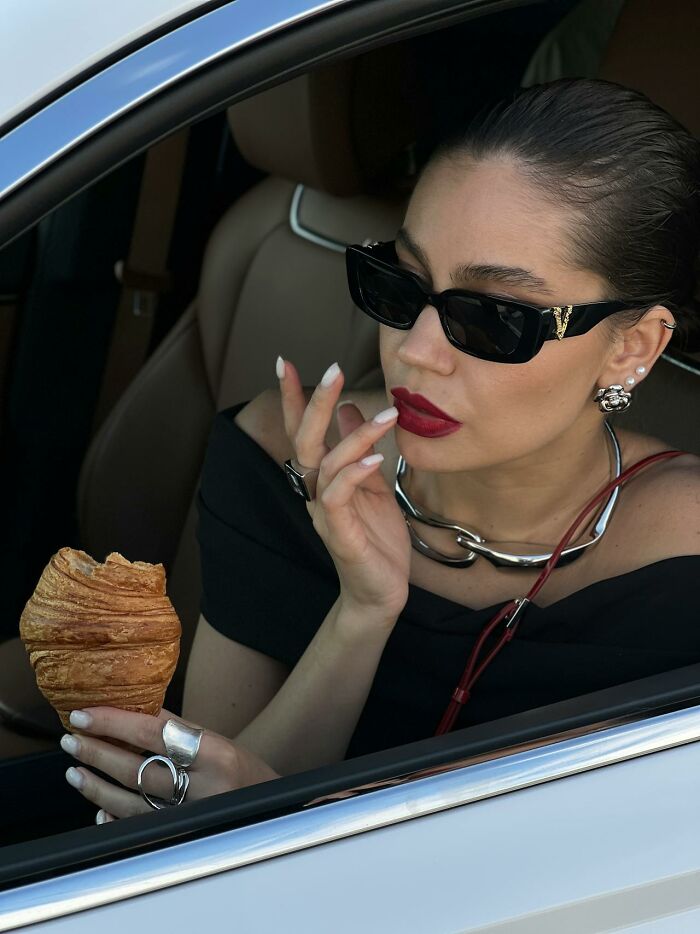 Woman in sunglasses holding a croissant, seated in a car, looking thoughtful.