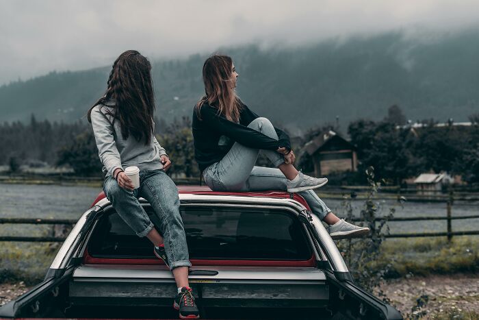 Two women sitting on a car by a lake, showcasing bisexual dating experiences in different gender contexts.
