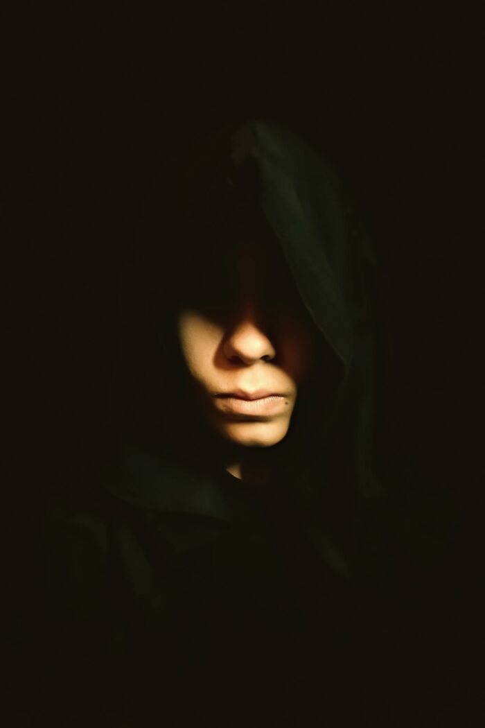 A person in a dark hooded cloak, casting shadows on their face, evoking a sense of scary true story atmosphere.