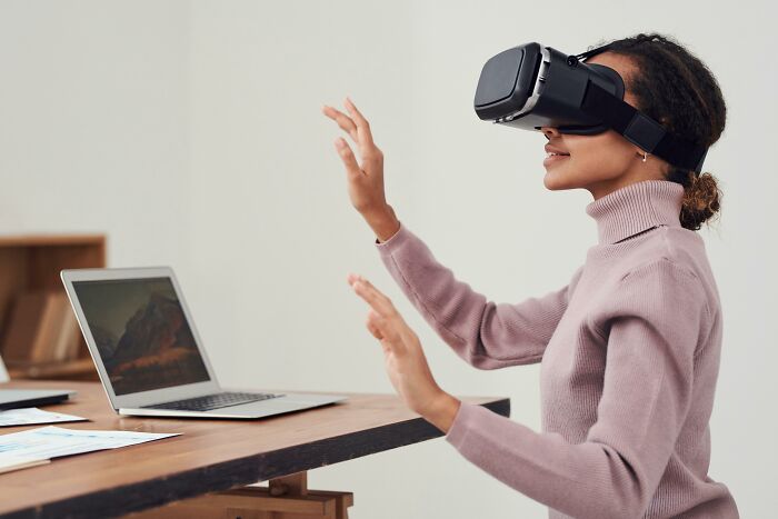 Person using VR headset at a desk with laptop, embodying office expressions with virtual gestures.