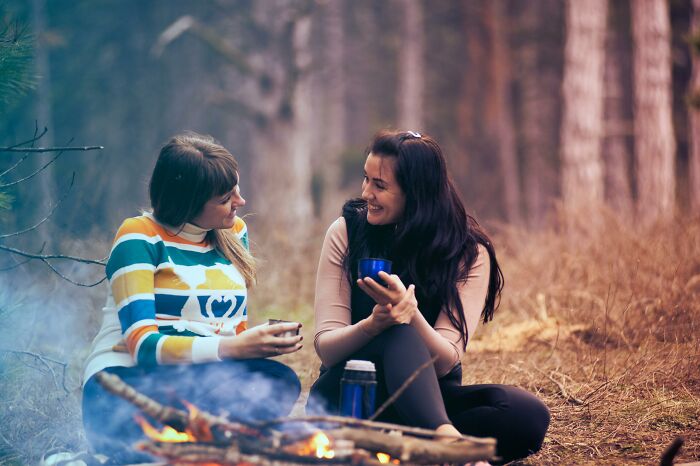 Two women in a thoughtful conversation by a campfire, holding mugs, surrounded by nature, showcasing intelligence.