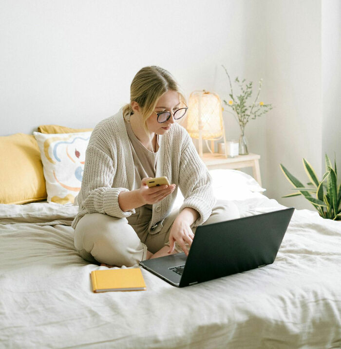 Person sitting on a bed, using a laptop and phone, possibly related to being fired by their boss.