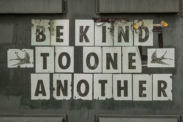 Sign with the message "Be Kind to One Another" on a textured wall, highlighting scams.