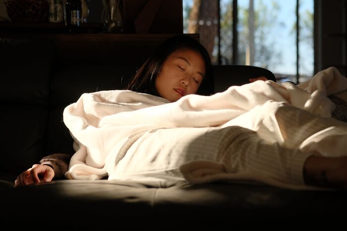 Woman sleeping on a couch wrapped in a blanket, with sunlight filtering through nearby windows.