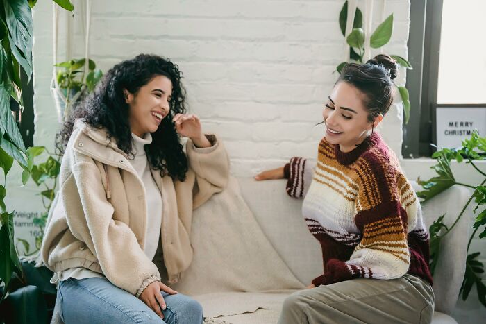 Two women sitting on a couch, sharing heartfelt compliments, surrounded by plants in a cozy room.