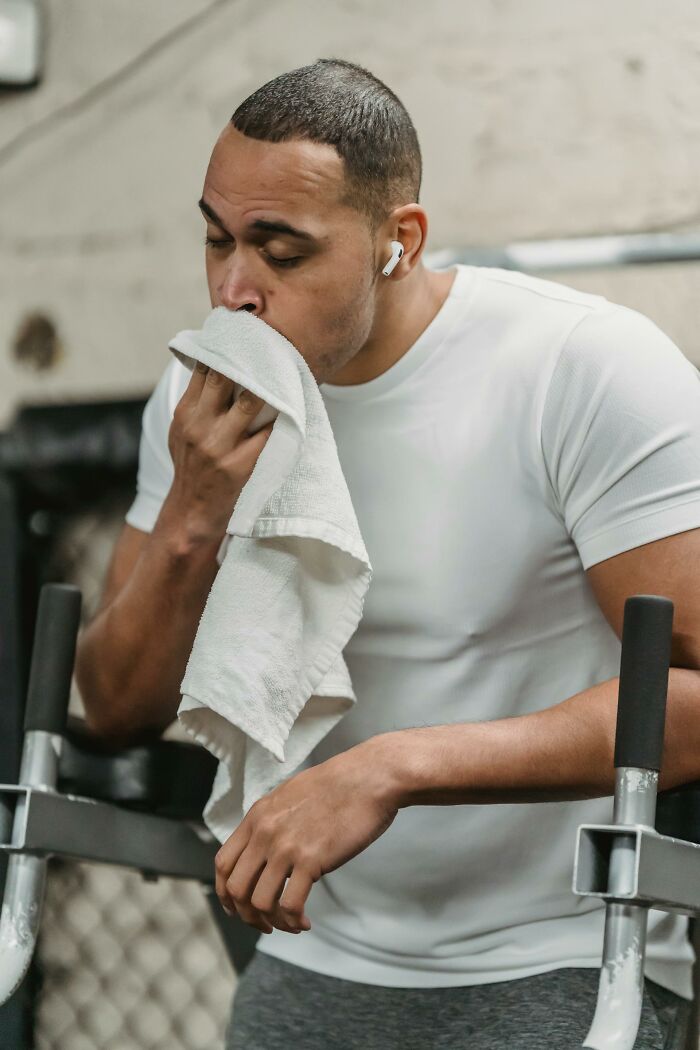 Man in gym wears earbuds, wipes face with towel, resting on exercise equipment.