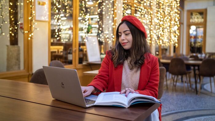 Woman in red coat and beret using a laptop at an outdoor cafe, lights twinkling.