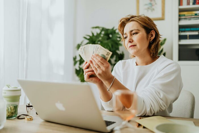 Person in white sweater holding cash, sitting at a desk with a laptop and cup, challenging stereotypes.