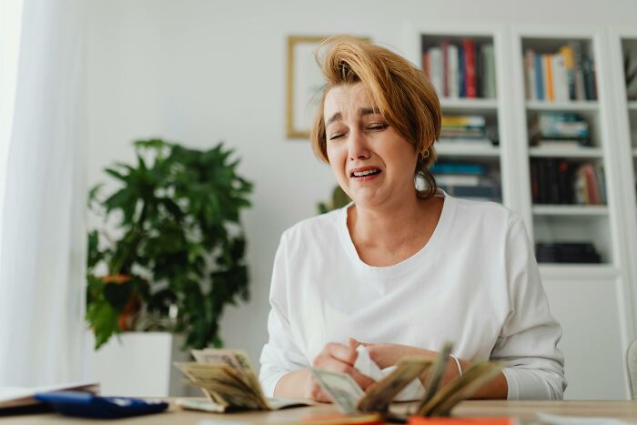 A woman in a white shirt at a table, counting money with an expression of emotional distress, reflecting weirdest thing society accepts as normal