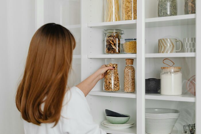 Woman organizing a middle-class home kitchen shelf with jars and dishes, focusing on practical storage solutions.