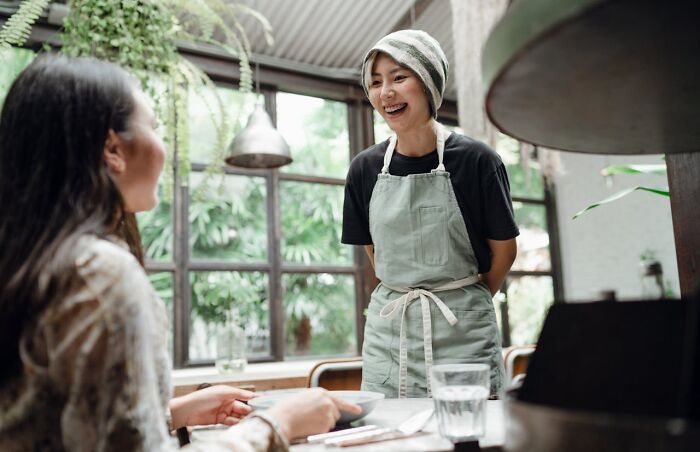 Woman in a green apron smiling and conversing with another woman in a peaceful, plant-filled cafe setting.