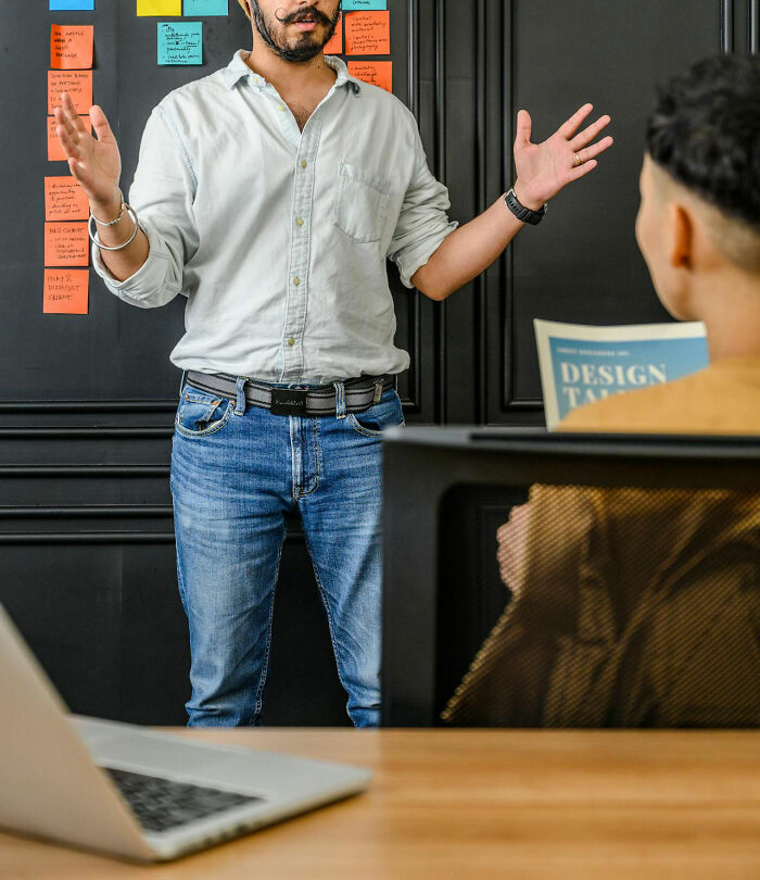 Person presenting in front of a blackboard with sticky notes, discussing a work-related issue in an office setting.