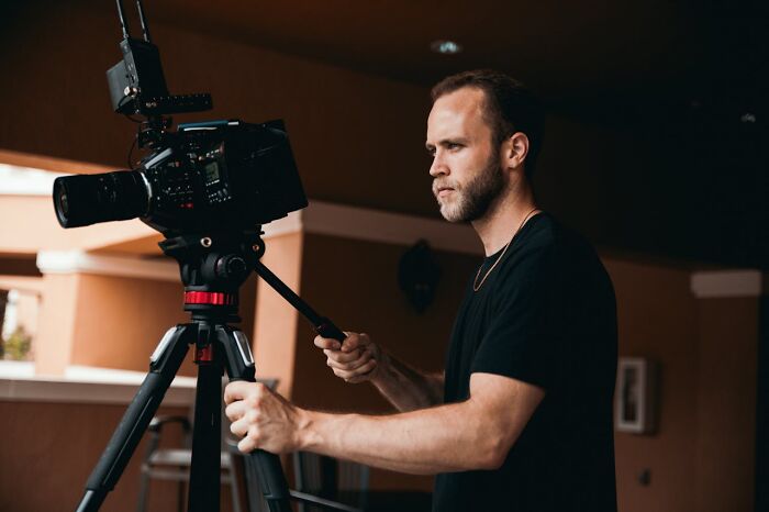 A man operating a professional video camera on a tripod in a dimly lit room.