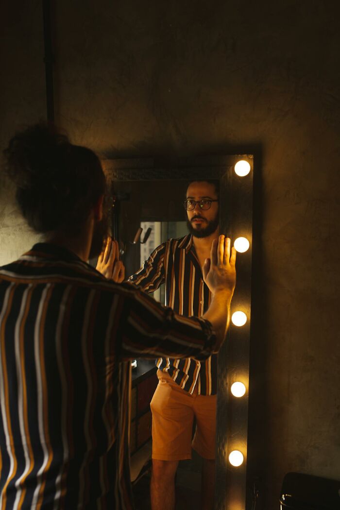 Man in striped shirt and glasses looks in a lit mirror, reflecting on personal moments of rejection.