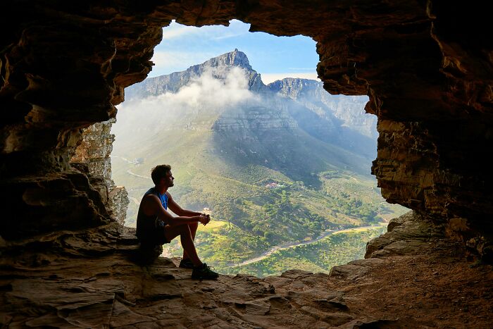 Man sitting in a cave, overlooking a breathtaking natural wonder landscape with mountains and clouds in the distance.