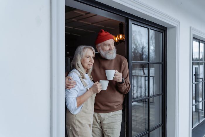 Elderly couple with mugs, overlooking a porch, sharing a moment of privacy.
