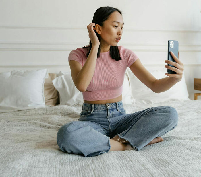 Woman in a pink top sitting on a bed, taking a selfie with her phone, reflecting on relationships.