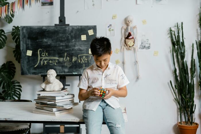 Young boy solving a Rubik's Cube, sitting by stacked books in a classroom, suggesting concepts of IQ and intelligence.