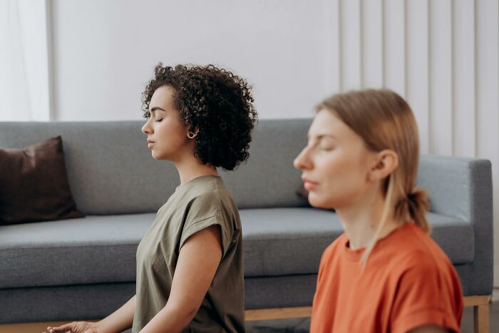 Two women meditating on the floor, a smart way to stay focused and mindful, seated in front of a sofa.