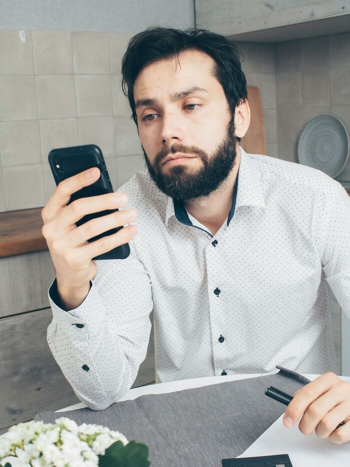Man in a white shirt looking at his phone thoughtfully, seated at a table, contemplating easy money-making ideas.
