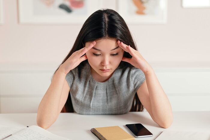A woman sitting at a desk, appearing stressed, with a notepad and smartphone, questioning societal norms.