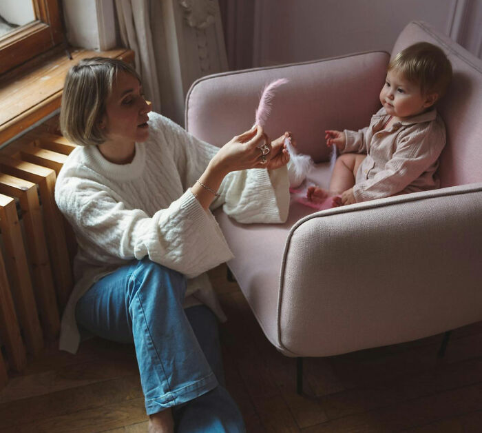 A woman in a cozy room playing with a pink plush toy with a baby in a chair.
