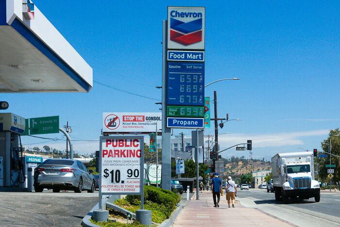 Chevron gas station signage showing high fuel prices, with a public parking sign in the foreground under a clear blue sky.