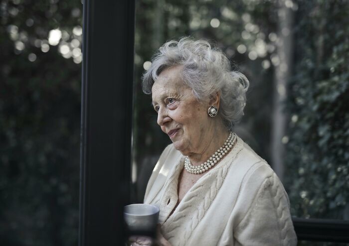 Elderly woman with white hair and pearl necklace, holding a cup, indoors with greenery visible through a window.