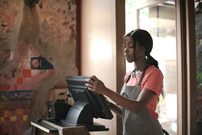 Woman in apron using a cash register, capturing a moment of a heartfelt compliment in a cozy, rustic cafe setting.
