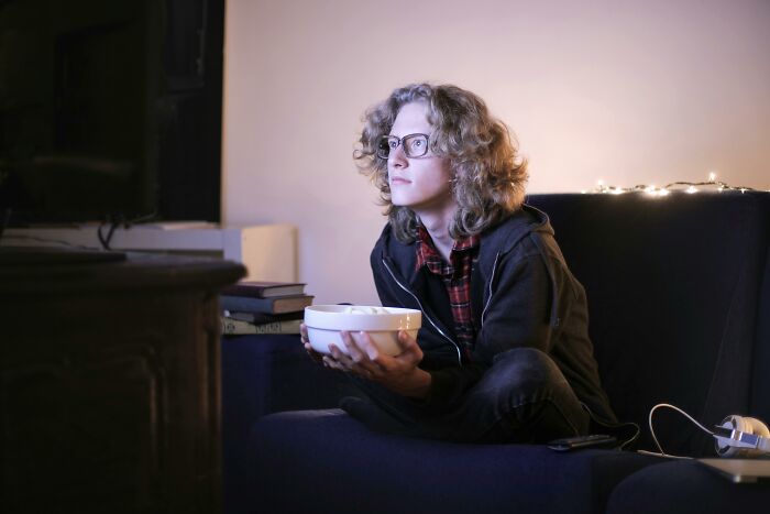 A person with glasses watching TV intently, glowing lights in the background, holding a bowl with snacks.