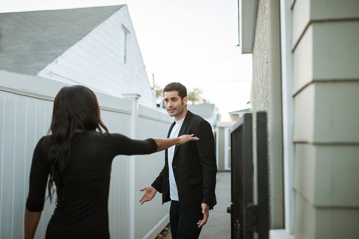 Person in a black jacket gesturing with open arms, facing a woman in a black dress in an alleyway, showing emotion.