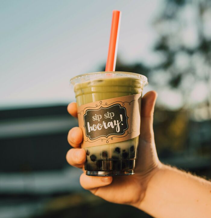 A hand holds a cup of bubble tea with a green matcha drink and tapioca pearls, labeled "sip sip hooray" in sunlight.