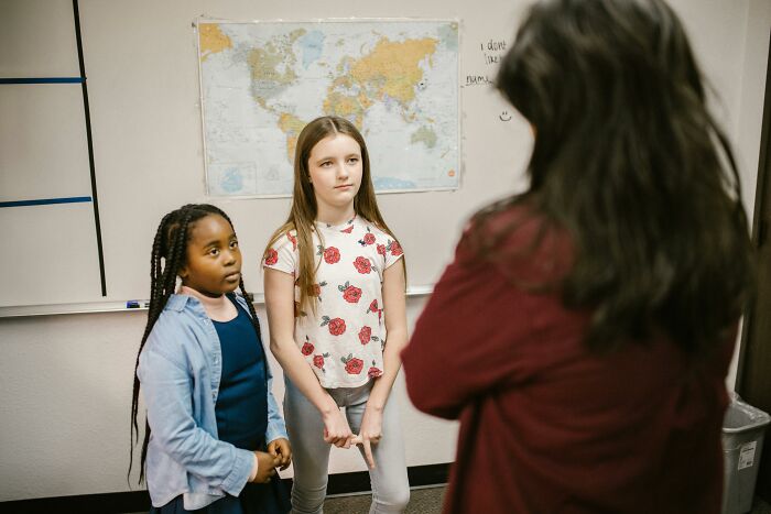 Two young students stand in a classroom, looking at a teacher, with a world map in the background.