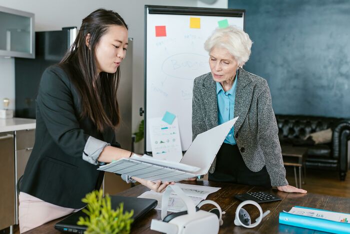 Two women in a business meeting, one reviewing documents, highlighting heartfelt compliments in a modern office space.