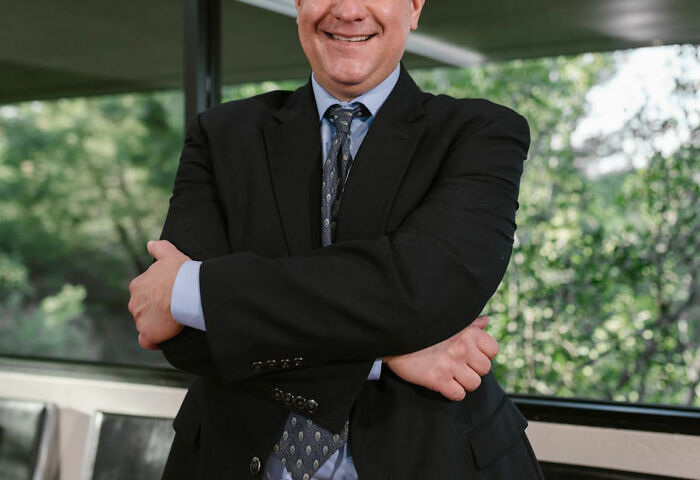 Man in a suit standing with arms crossed by a window, smiling confidently; represents bosses and employee dynamics.