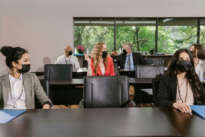People in a meeting room wearing masks and varied professional attire, chatting, with a green landscape view outside the window.