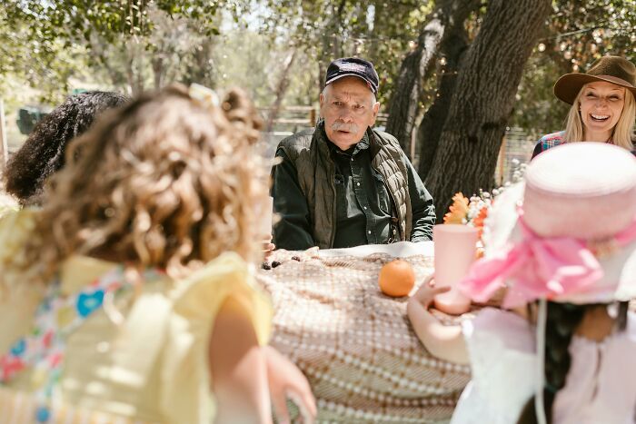 Older man at a picnic table receiving heartfelt compliments from children dressed for a tea party.