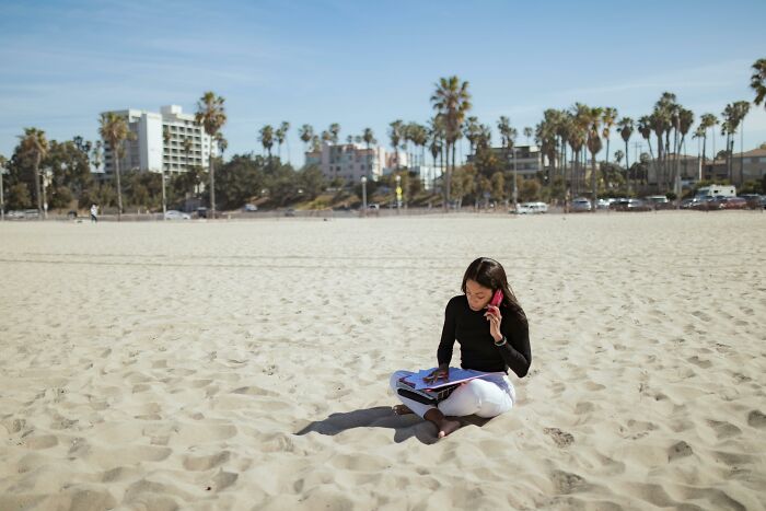 Person on a sunny beach using a phone and reading, surrounded by palm trees and buildings.