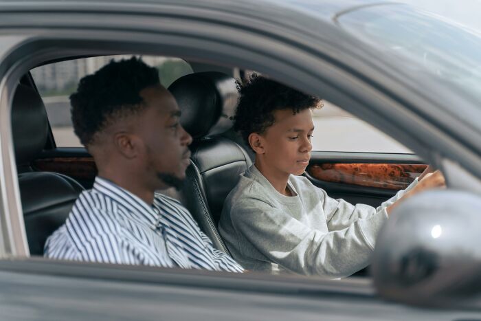 A young boy in a car tries driving with an older male passenger, sharing a moment over an insignificant thing.