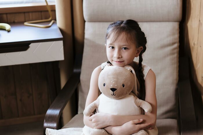 A young girl with braids hugs a plush bear, sitting in a cozy chair, embodying a heartfelt moment.