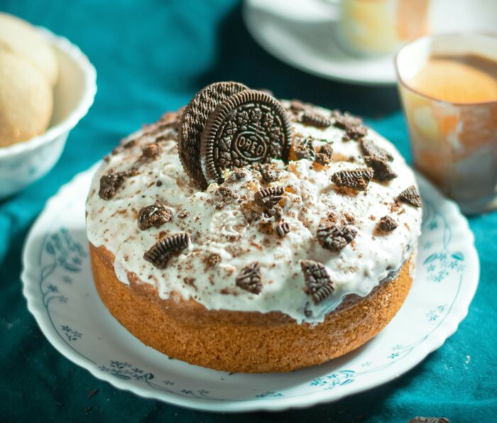 Oreo cookie cake with white frosting on a decorative plate, surrounded by other foods and a beverage.