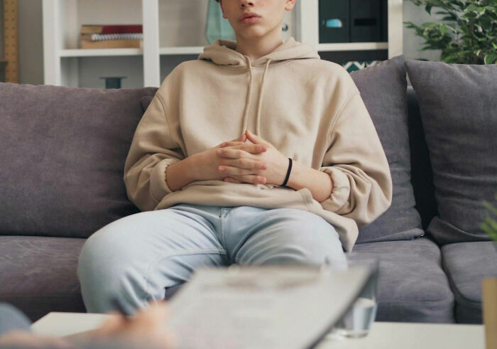 Person in a beige hoodie sitting on a couch during a therapy session, with a therapist holding notes in the foreground.