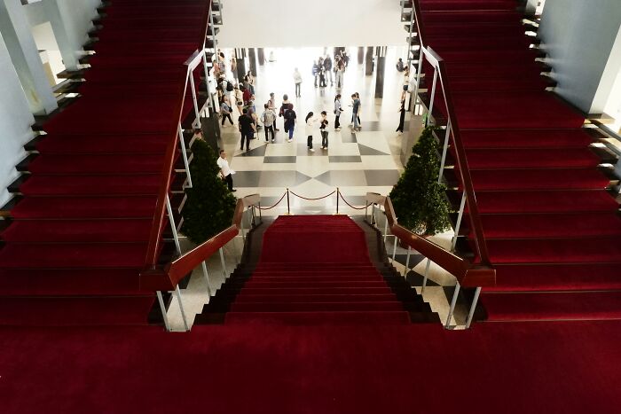 Red carpeted stairs leading to an open lobby with people gathered, hinting at celebrity conspiracy themes.